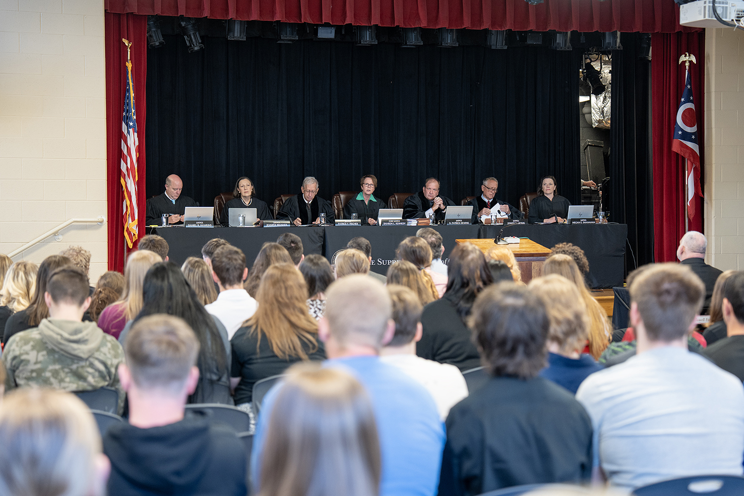 A long table at which are seated seven men and women all wearing black judicial robes in front of an audience of high school students. 