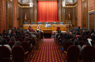 Two women are seated in red, velvet chairs to the right of another woman speaking from a wooden podium to an audience of men and women in the courtroom of the Thomas J. Moyer Ohio Judicial Center.