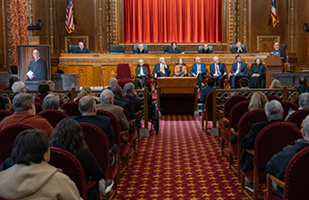 A man speaks from a wooden podium to a large group of men and women in the courtroom of the Thomas J. Moyer Ohio Judicial Center. On his right, seven men and women are seated in red, velvet chairs. To their right is a portrait of the man speaking from the podium. Behind them are five men and women all wearing black judicial robes seated at a wooden bench.