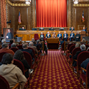 A man speaks from a wooden podium to a large group of men and women in the courtroom of the Thomas J. Moyer Ohio Judicial Center. On his right, seven men and women are seated in red, velvet chairs. To their right is a portrait of the man speaking from the podium. Behind them are five men and women all wearing black judicial robes seated at a wooden bench.