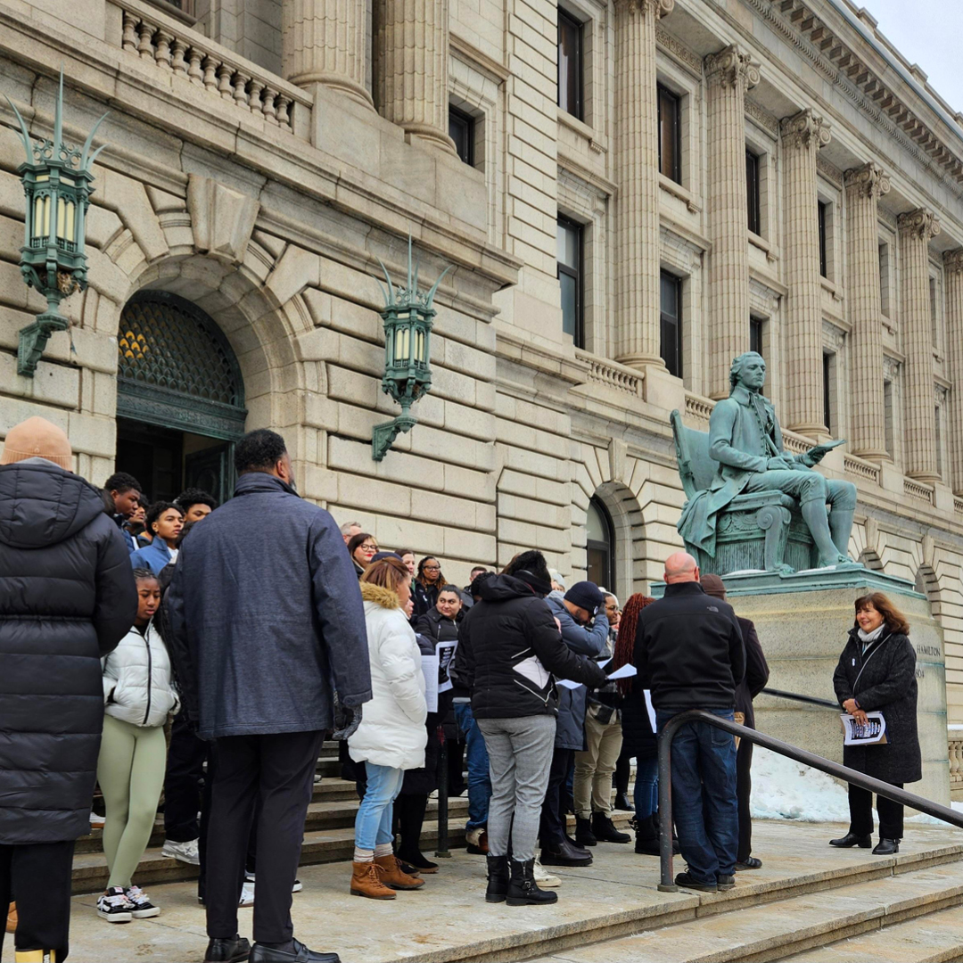 A group of men and women wearing winter coats standing outside listening to a woman speak with a booklet