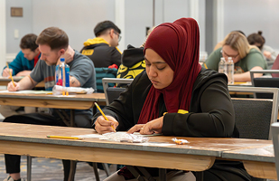 A woman wearing a maroon head scarf and black blouse sits at a long, narrow wooden writing on a paper with a yellow pencil.