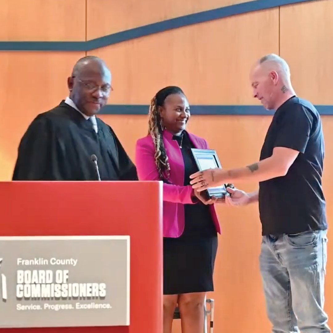 A judge and a woman in a pink blazer handing an award to a man in a black shirt