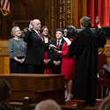 A man raising his right hand on a bible a woman reading from a book holding up her right hand and an audience watching around them.