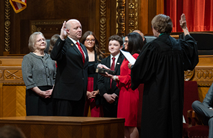 A man raising his right hand on a bible a woman reading from a book holding up her right hand and an audience watching around them