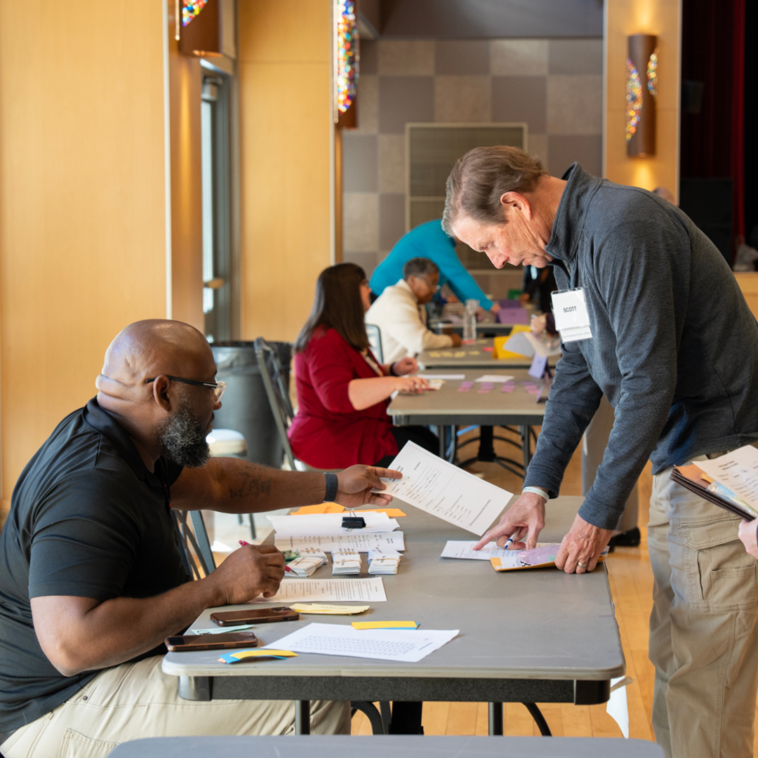 A man sitting at a table showing a man standing documents in a large hall