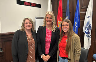 A woman wearing a brown polka dot blouse and black blazer, a woman wearing a bright pink polka dot blouse and black blazer, and a woman wearing jeans, a red shirt, and brown jacket standing side-by-side.