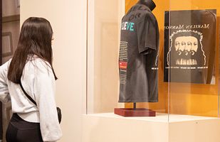 A young woman wearing black yoga pants and a white sweatshirt looks at a black t-shirt on display in a plexiglass case.