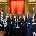 Nine students and three teachers smiling with a trophy in a courtroom