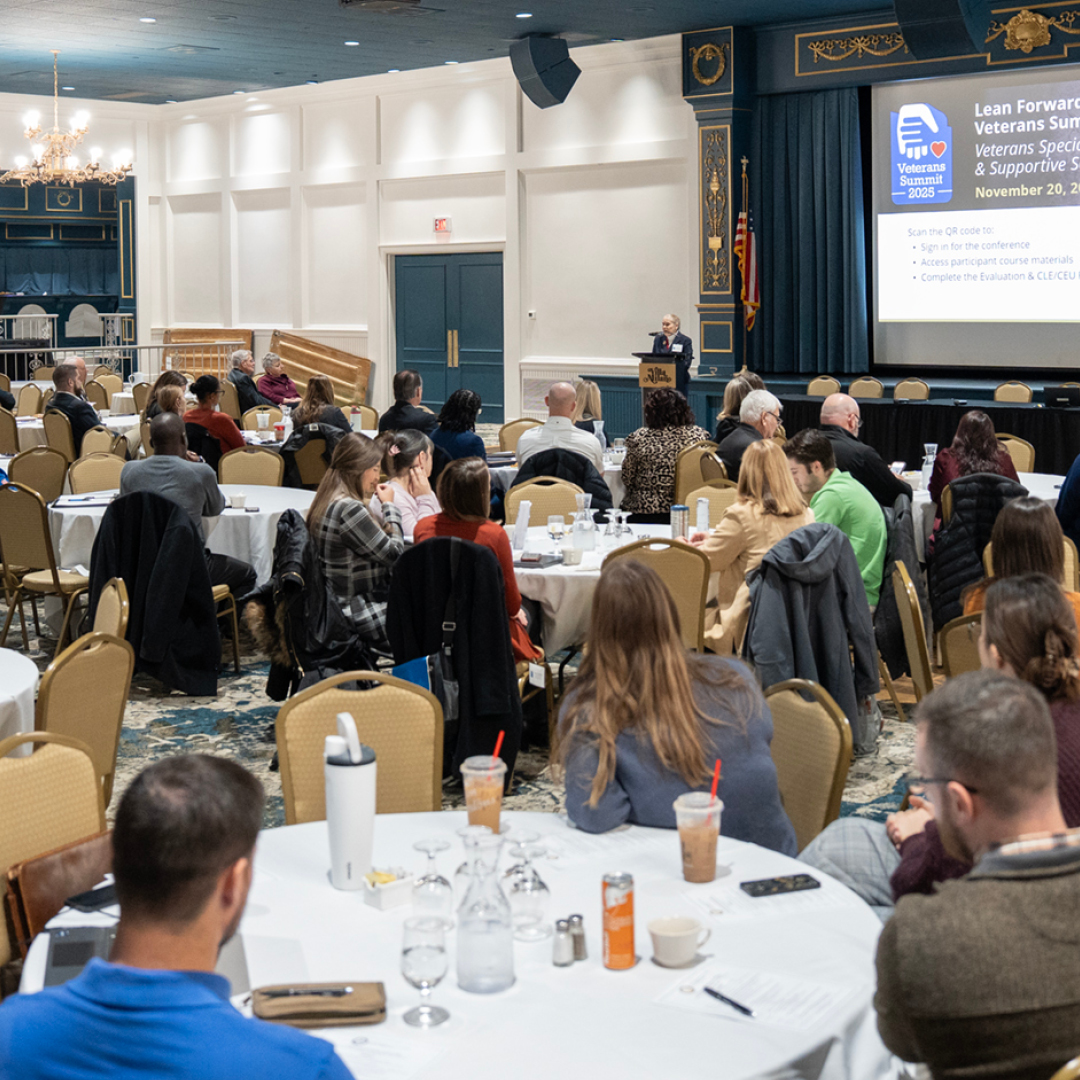 A woman speaking at a podiem with the US flag and Ohio flag in the background with a table of women listening to the speaker