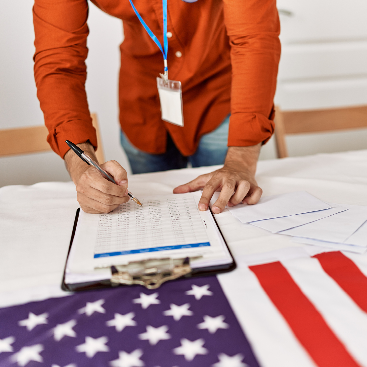Person in an orange shirt writes on a clipboard beside loose papers and an American flag.
