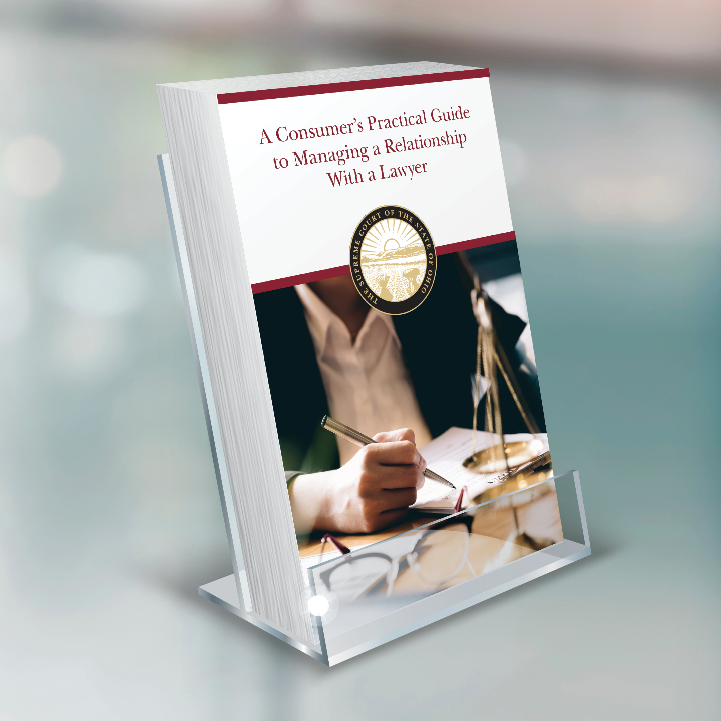 A book titled 'A Consumer’s Practical Guide to Managing a Relationship With a Lawyer' displayed in an acrylic stand, with a blurred background. A person is writing on papers with a pen, and eyeglasses are on the table.