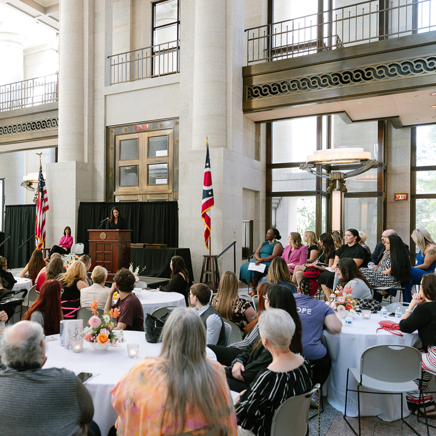 Speaker at a podium addresses a seated audience at a formal event in a bright stone atrium decorated with U.S. flags and flowers.