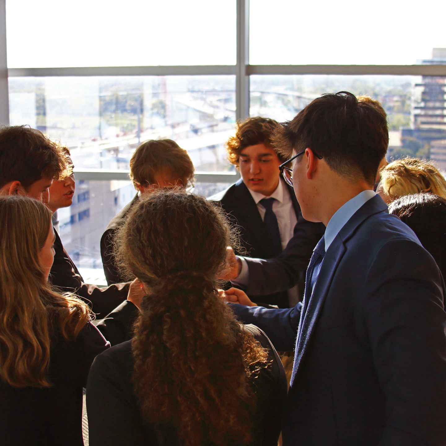 Group of formally dressed young adults talking near large windows overlooking a city.