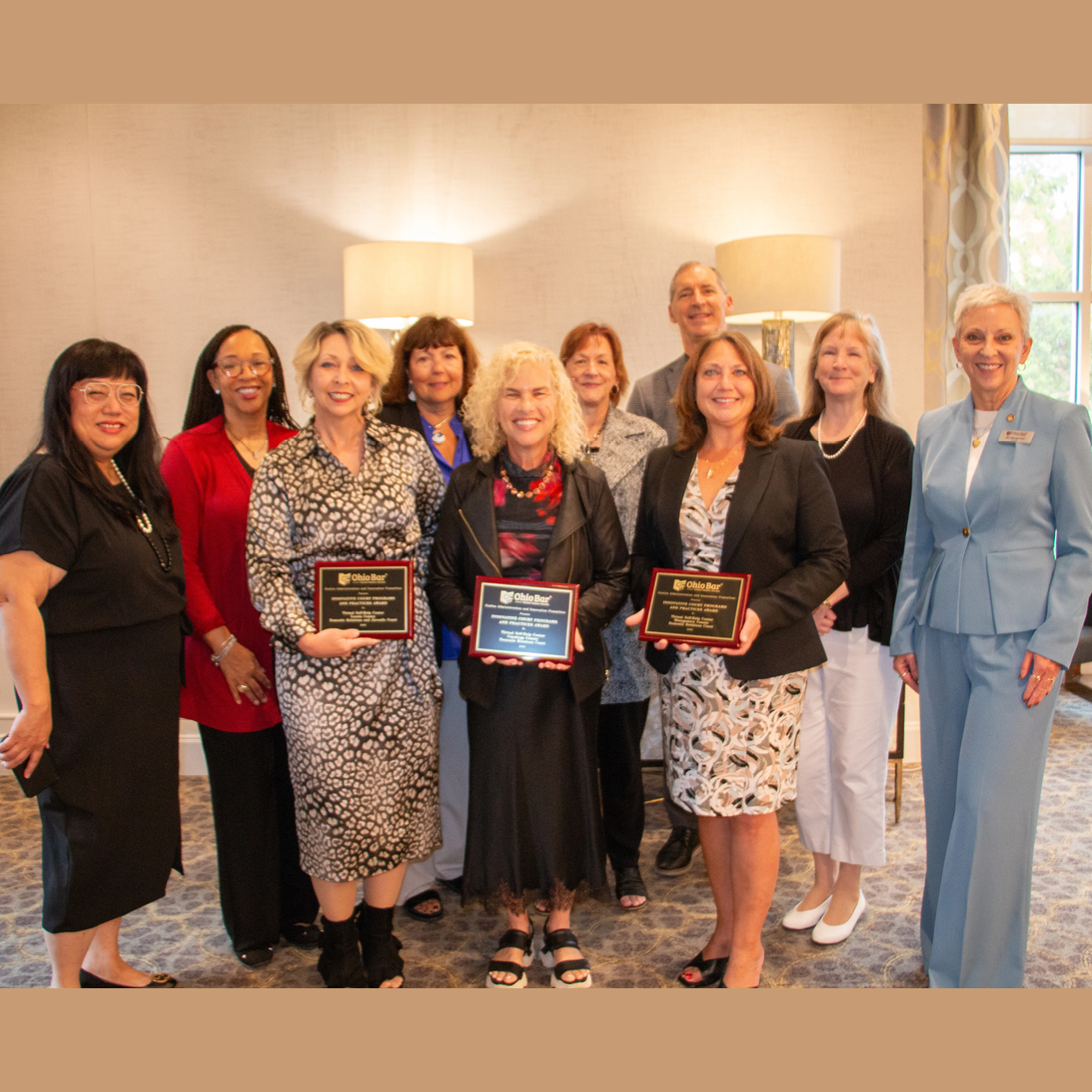 A group photo of ten professionals smiling together indoors, with three women in the front row holding wooden award plaques from the 'Ohio Bar.' The group features diverse attire, including formal business suits, patterned dresses, and casual cardigans, standing closely together in a well-lit room with large lamps and a window in the background.