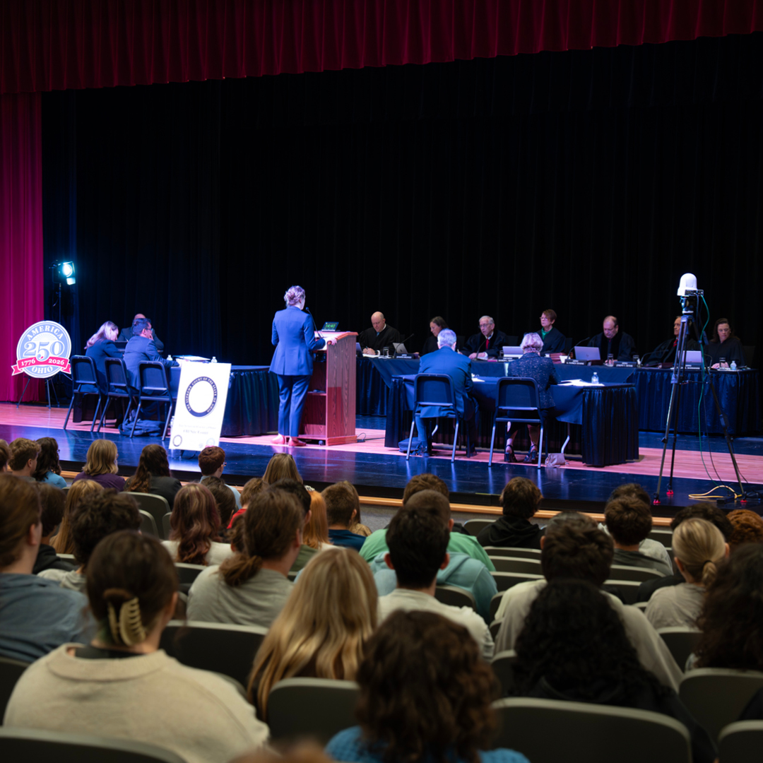 A courtroom scene on a stage with a person standing at a podium facing a panel of judges. An audience watches attentively from rows of seats.
