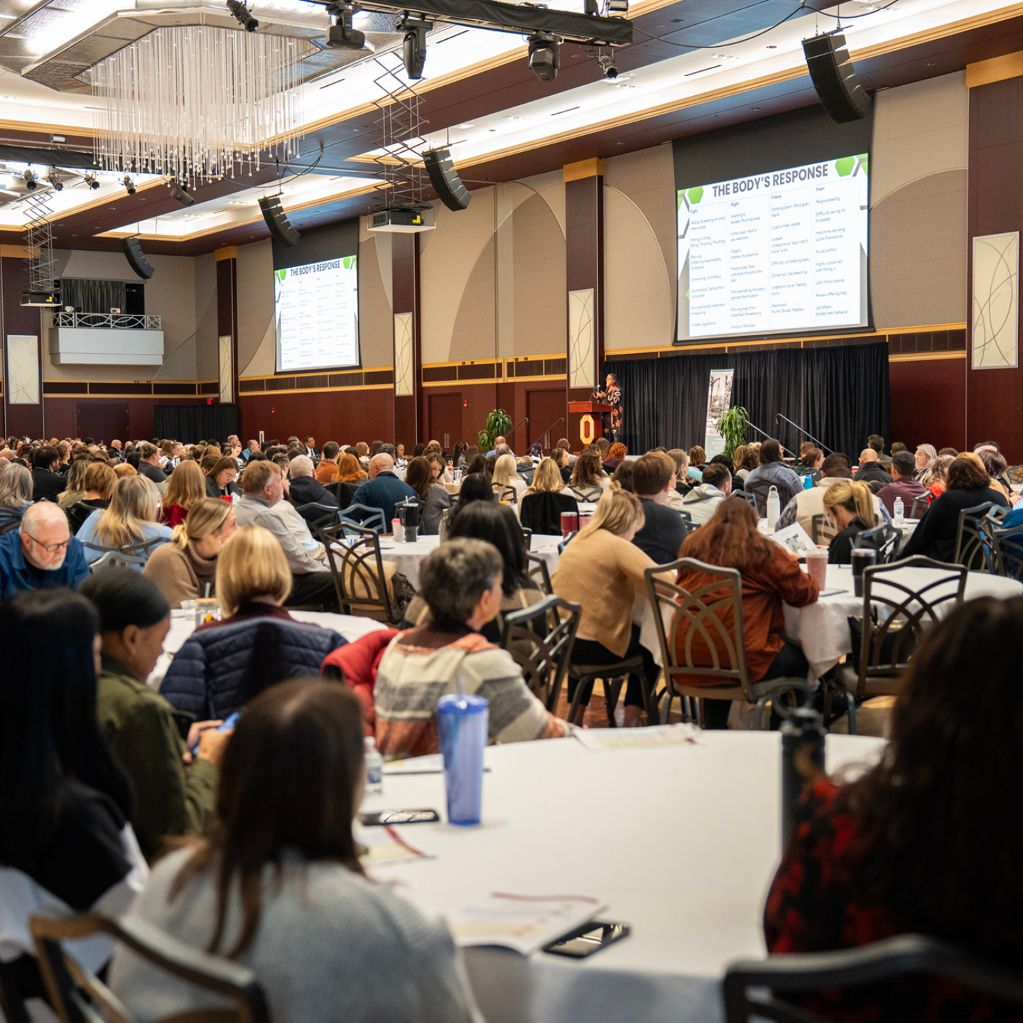 Speaker addressing a crowded conference room while 'THE BODY’S RESPONSE' appears on two projection screens.