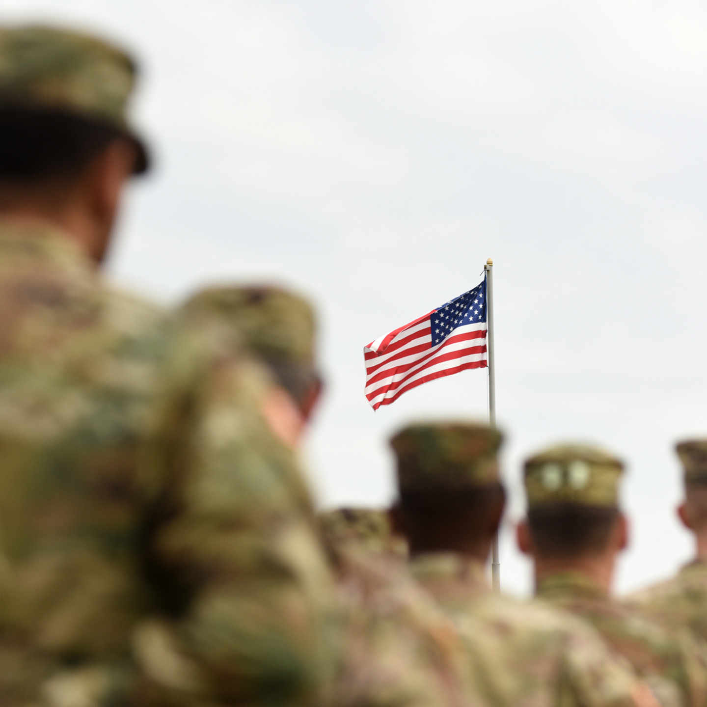 U.S. soldiers in uniform stand in formation with an American flag flying in the background on a cloudy day.