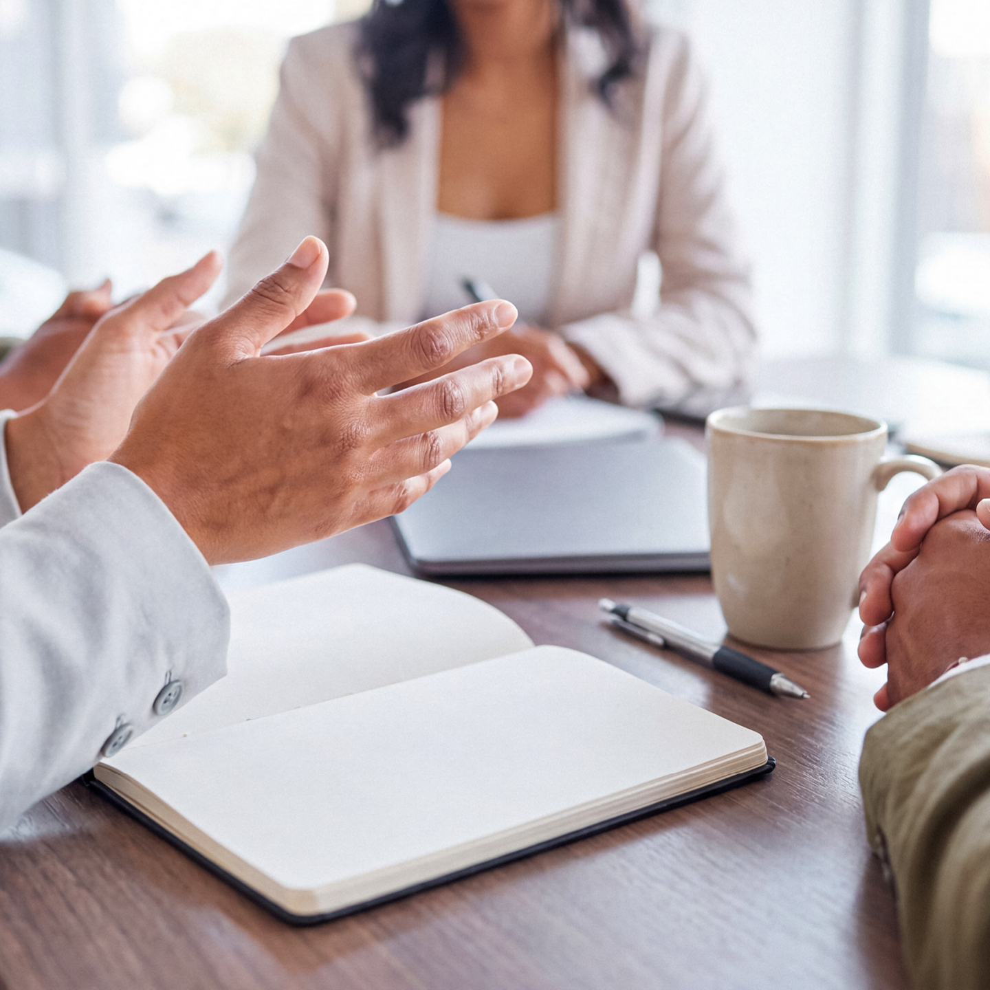 A close-up of a business meeting shows hands gesturing over an open notebook, a pen, and a coffee cup, suggesting active discussion and collaboration.