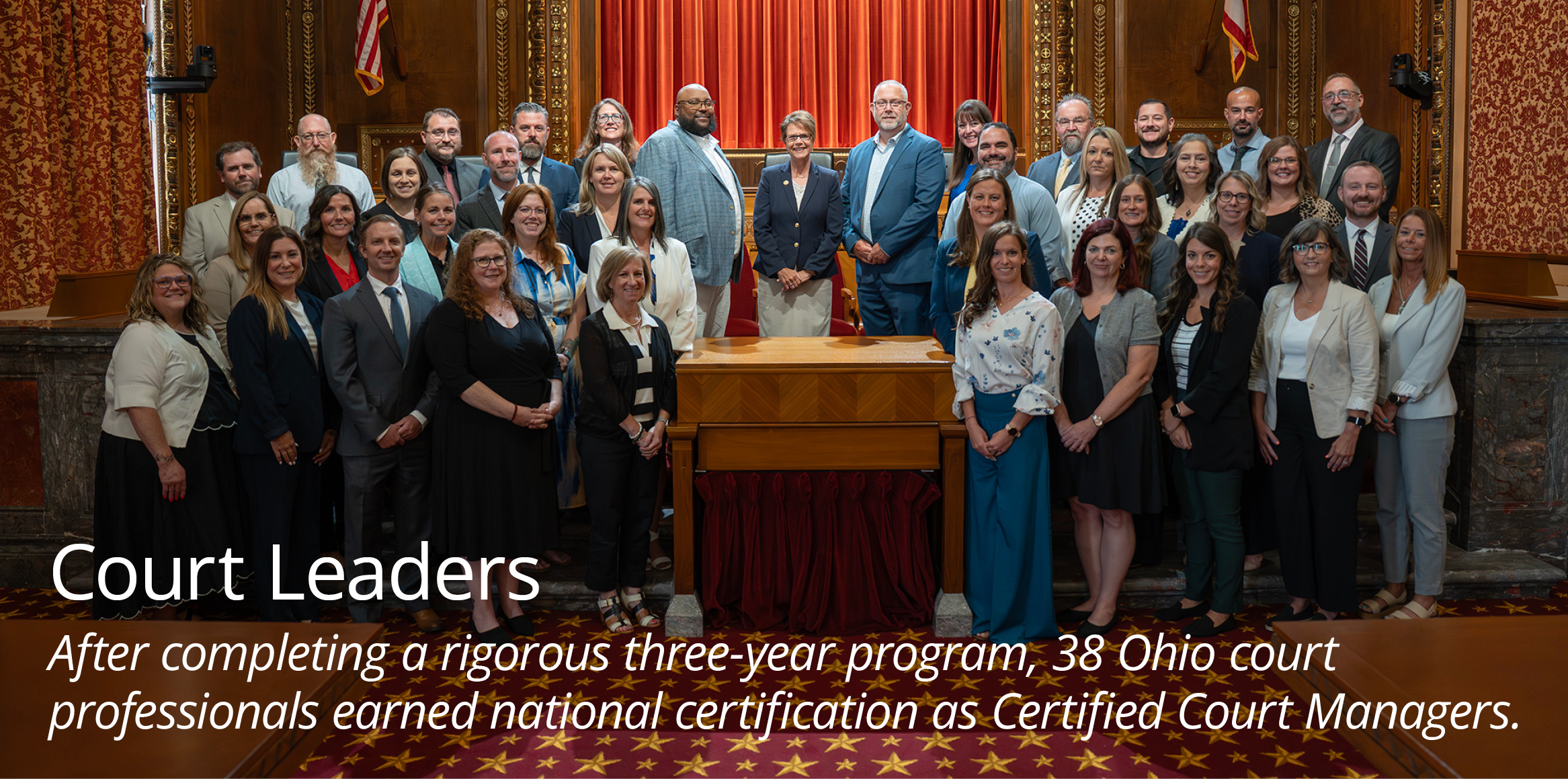 A large group of professionally dressed men and women pose for a group photo inside a grand, wood-paneled courtroom, standing in front of a wooden bench with a red curtain backdrop and the Ohio state flag.