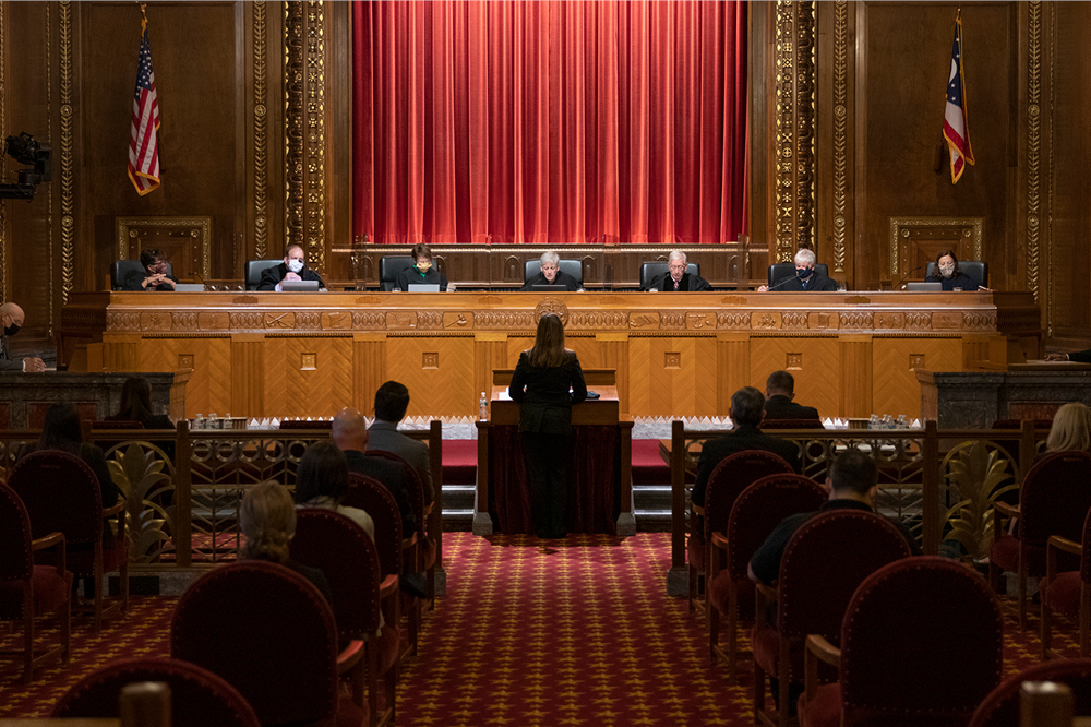 Image of a woman in a suit stading at a podium facing a bench of seven justices in the courtroom of the Thomas J. Moyer Ohio Judicial Center