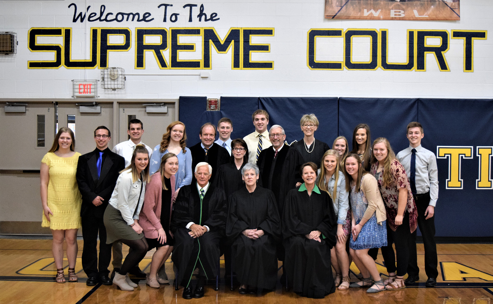 Image of seven justices wearing their black judicial robes posing with a group of high school students in a gymnasium