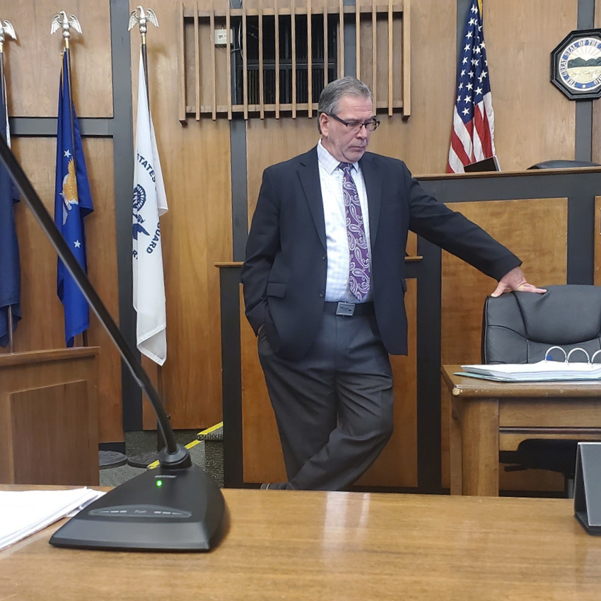 Image of a man wearing a dark suit and purple tie standing in a courtroom with his hand on a black office chair