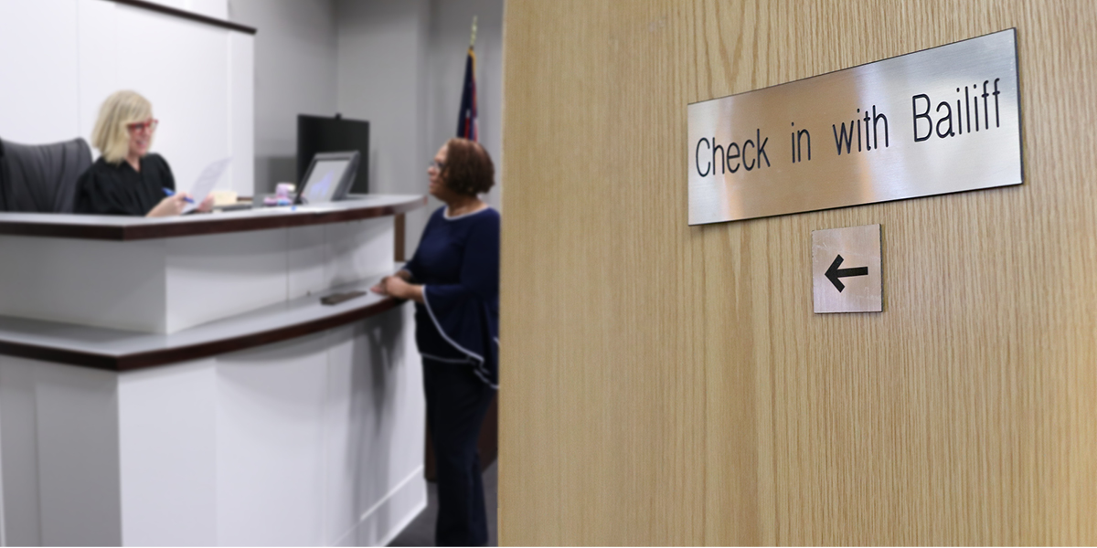 Image of an open wooden door with a sign on it that reads: 'Check in with Bailiff'. Beyond the door is a female bailiff talking with a female judge.
