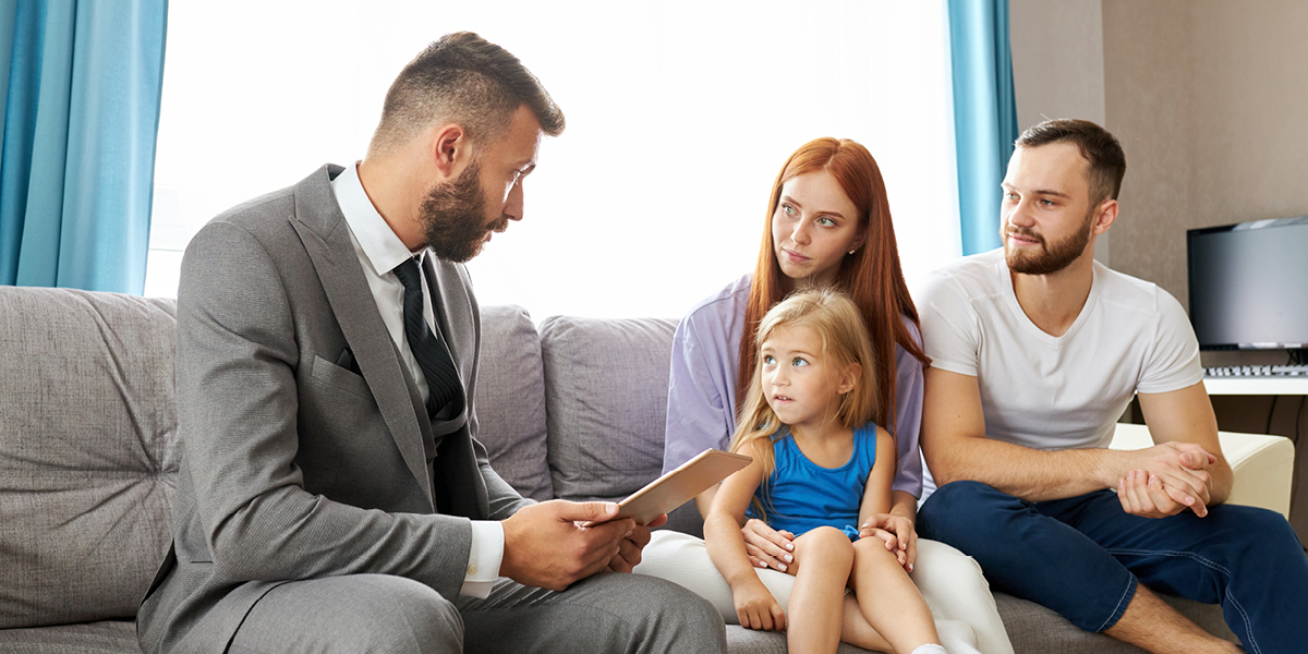 Image of a woman with long red hair holding a female child on her lap. Next to her is a man wearing a white t-shirt and blue pants. They are interacting with another man wearing a grey suit. All are sitting on a couch.