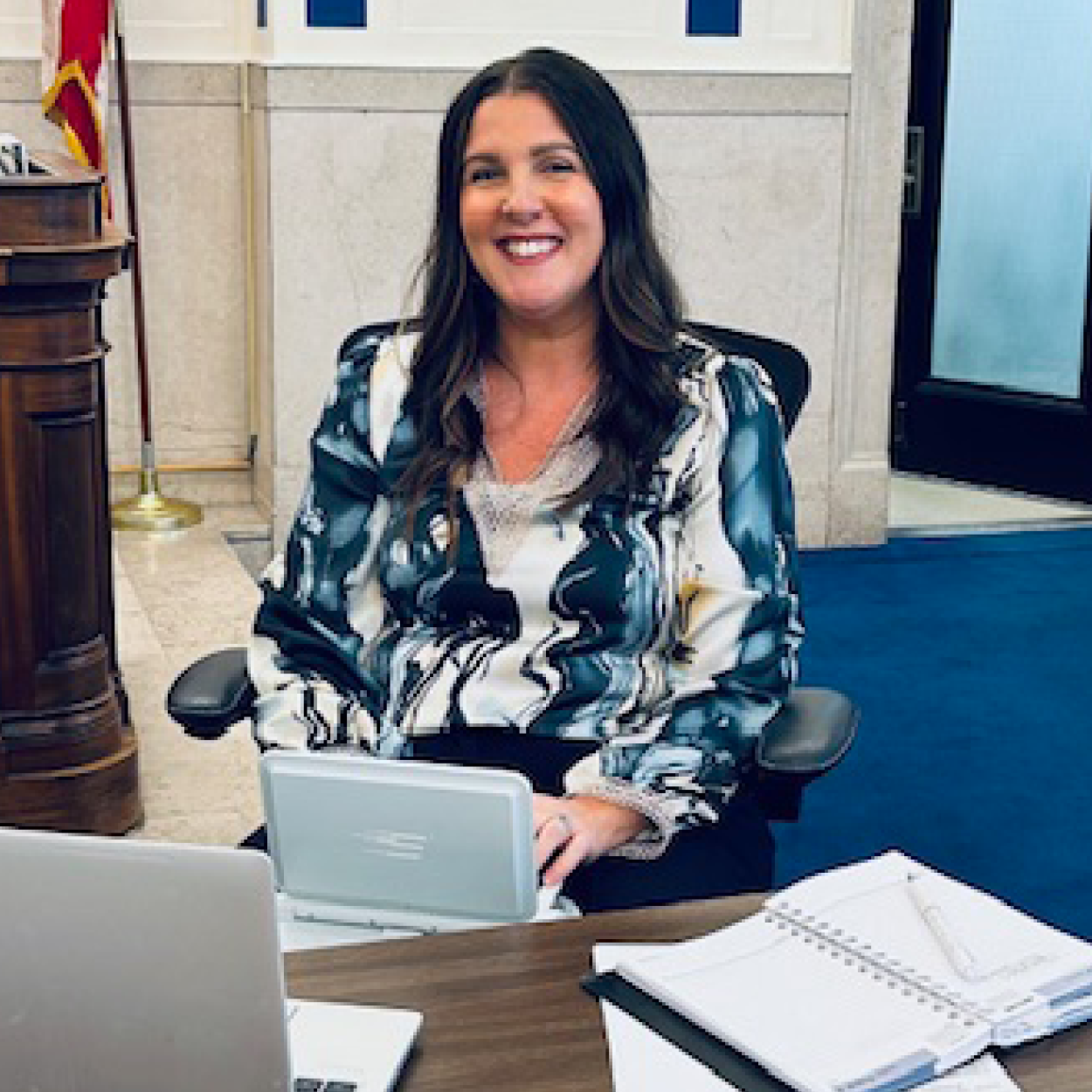 Image of a smiling woman with long, dark hair, wearing a blue and white patterned blouse, sitting at a desk in a courtroom.