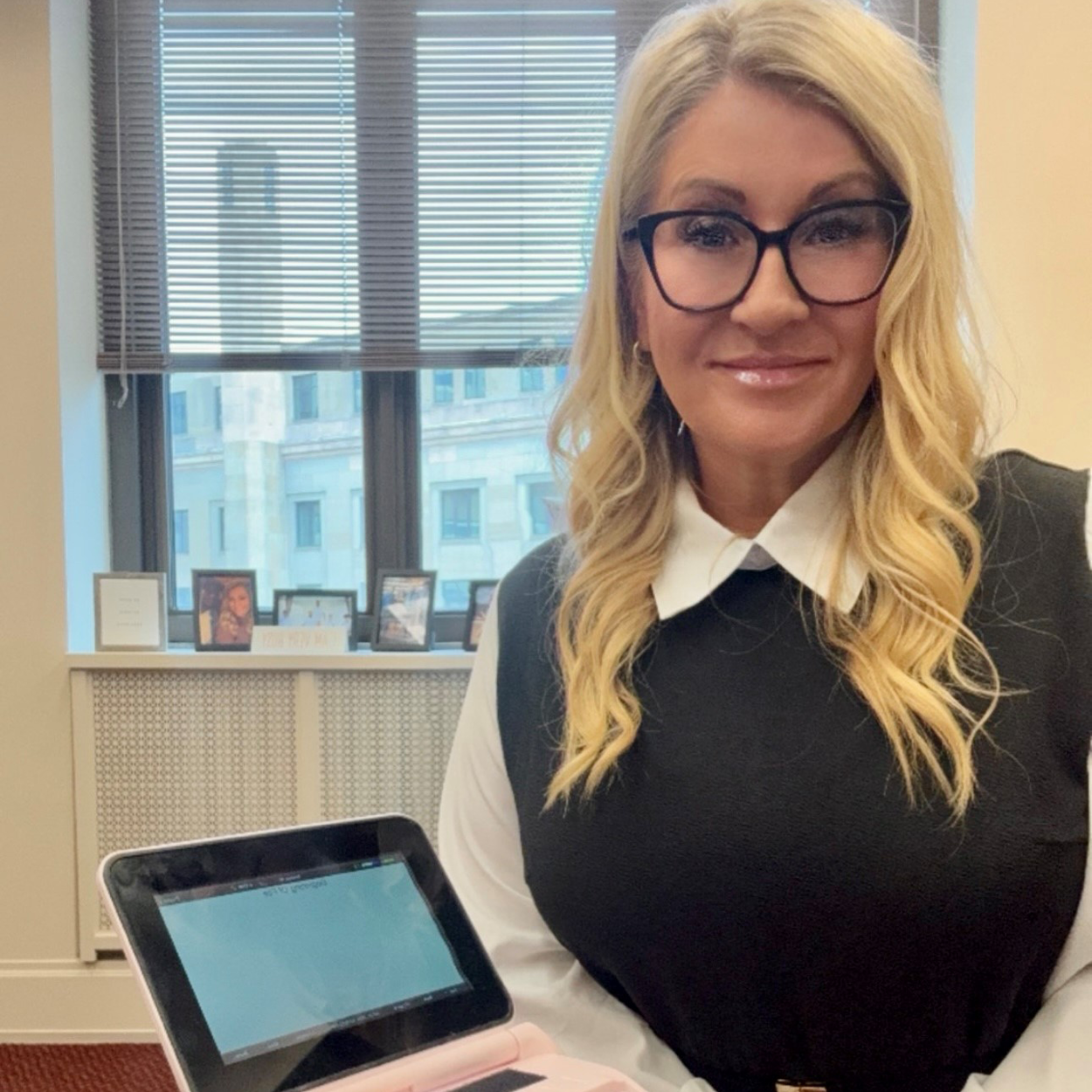 Image of a woman with long, blonde hair, wearing dark-rimmed glasses, a white blouse, and a black vest.