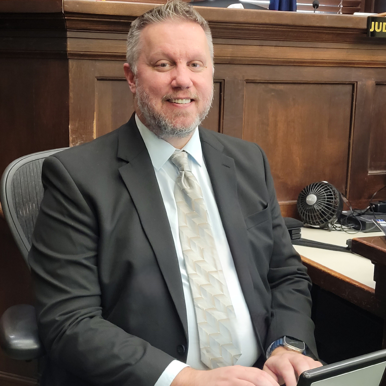 Image of a man wearing a grey suit, sitting in a chair in front of a stenotype machine in a courtroom.