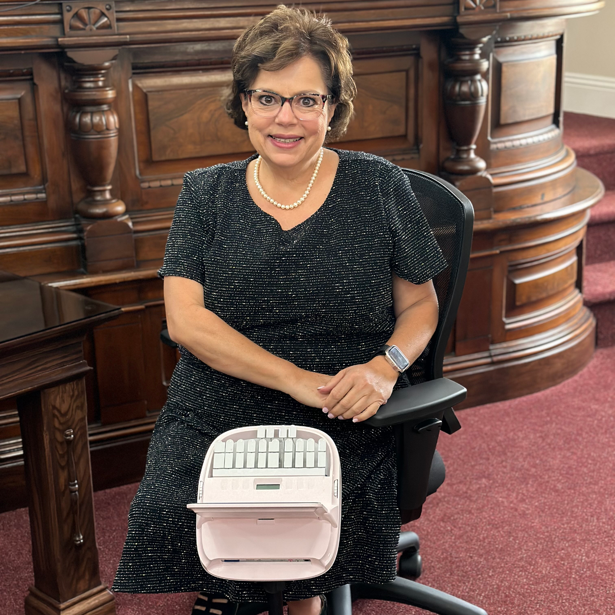 Image of a woman with short, dark hair and glasses, wearing a dark-colored dress, sitting in a courtroom behind a stenotype machine.