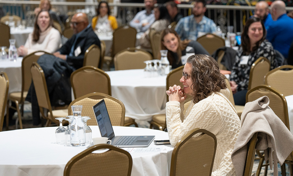 A woman wearing a white cable knit sweater sits alone at a round table with a white table cloth. In front of her is a laptop. She has her hands clasped and resting on her chin and she is smiling.