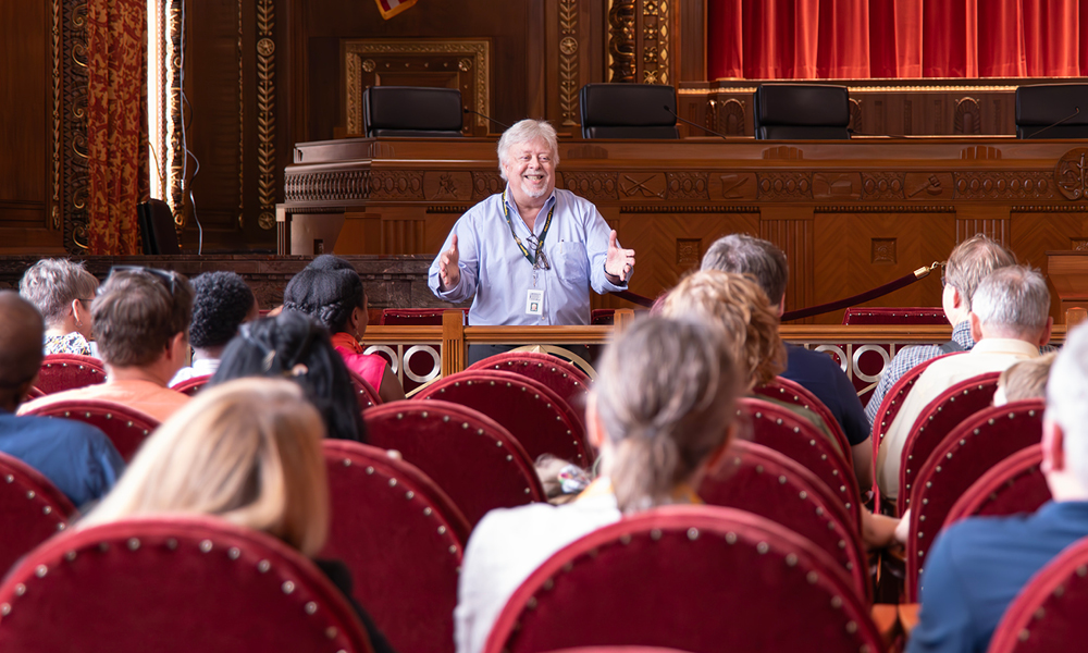 A man smiles and gestures to men and women seated in red velvet chairs in the courtroom of the Thomas J. Moyer Ohio Judicial Center.