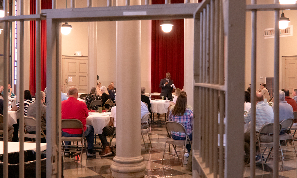 A man wearing a black suit and holding a microphone speaks to a group of men and women seated at round tables. The image is seen through an open jail cell door.