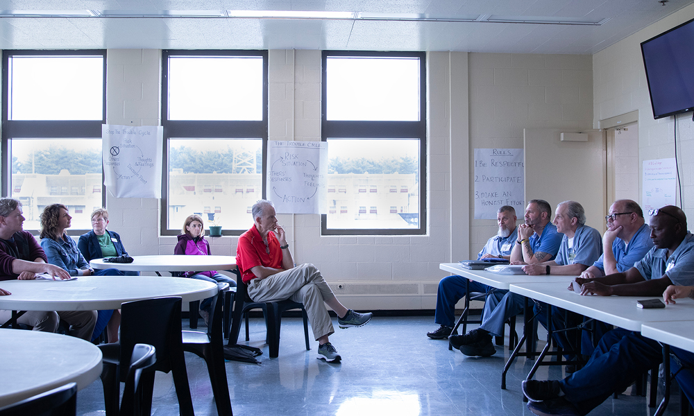 A man wearing a red, short-sleeve shirt sits with his legs crossed at a table with four women all listening to a group of men wearing blue prison clothes seated at long, white folding tables.