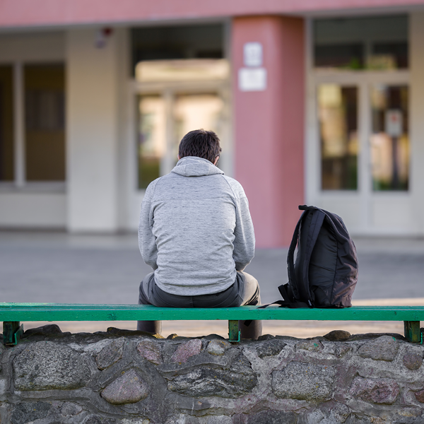 Image of a boy sitting next to a black bookbag on a green wooden bench with his back to the camera.