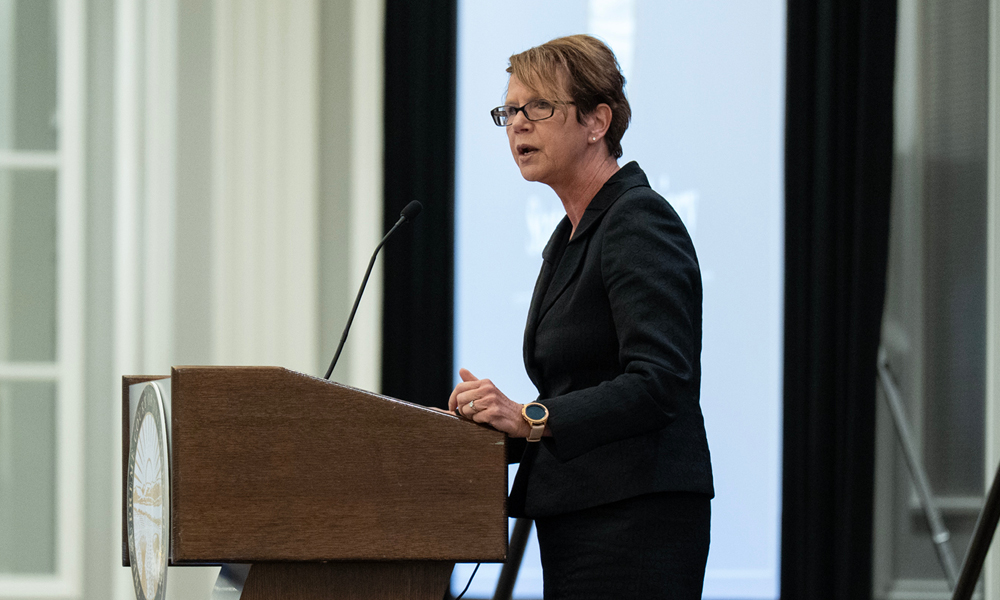 A woman wearing a dark suit speaks from a wooden podium.