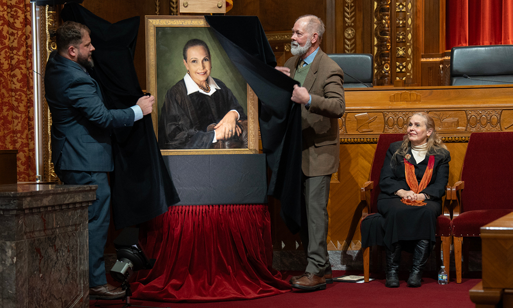 Two men remove a black cloth cover revealing a painted portrait of a woman wearing a black judicial robe. The woman in the portrait is seated in a chair beside the portrait.